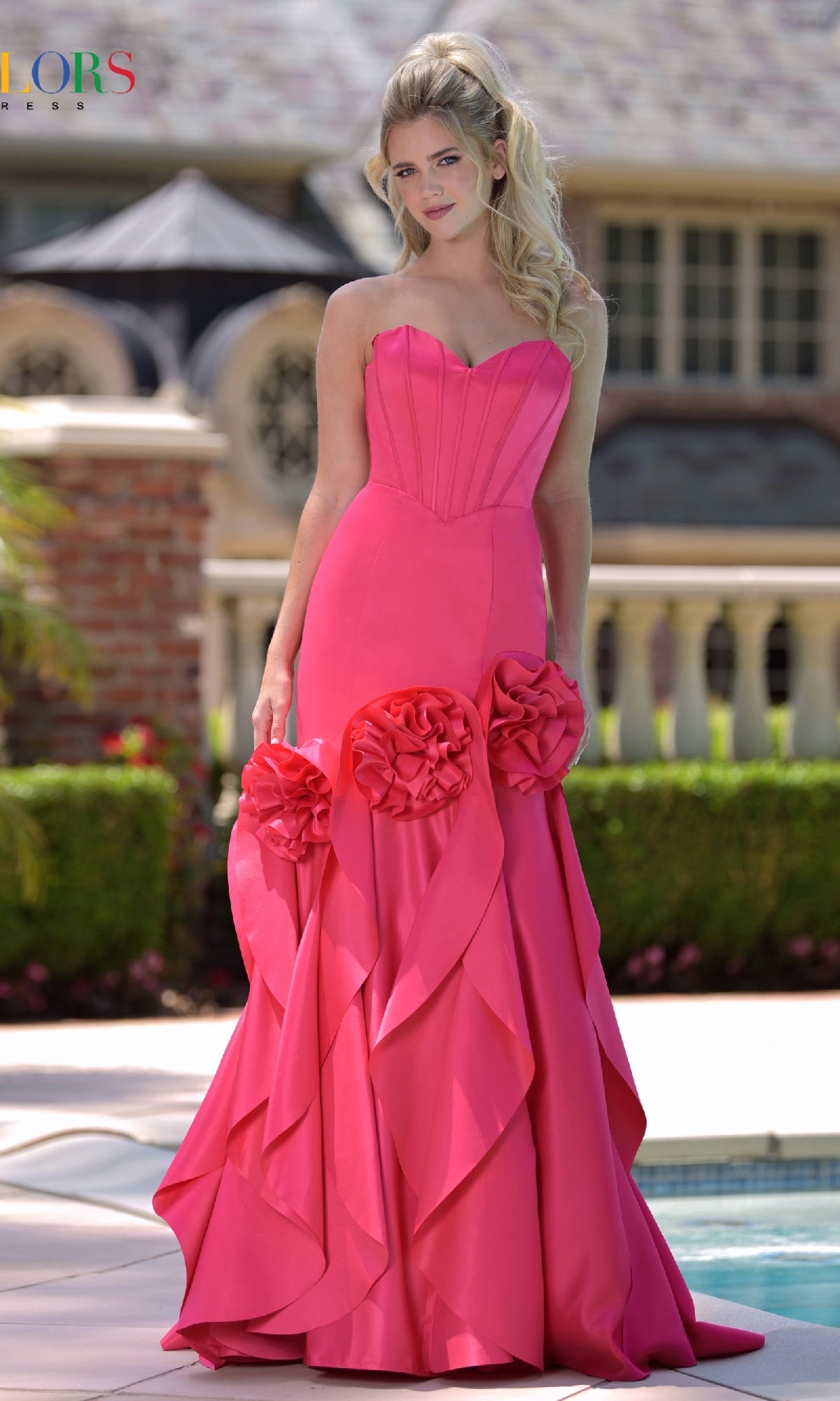 A woman stands outdoors by a pool, wearing the Colors Dress 3880— a strapless long prom dress featuring a corset bodice and large 3D rosette appliques on the skirt, with a building visible in the background.